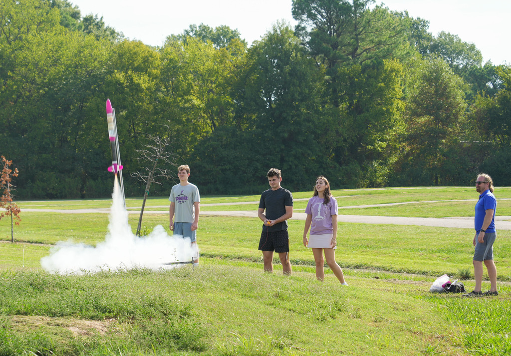 A rocket launching on a grassy field, with four people standing nearby. Trees and shrubs are in the background.