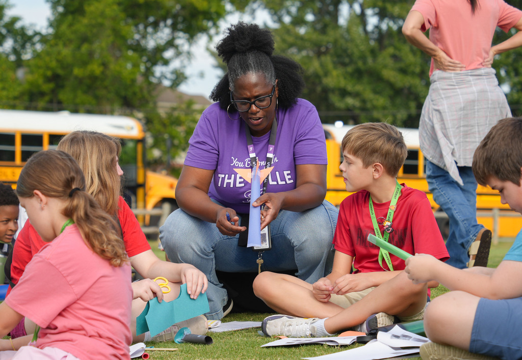 A woman in a purple shirt kneels, surrounded by children in casual clothes, all seated on a lawn.