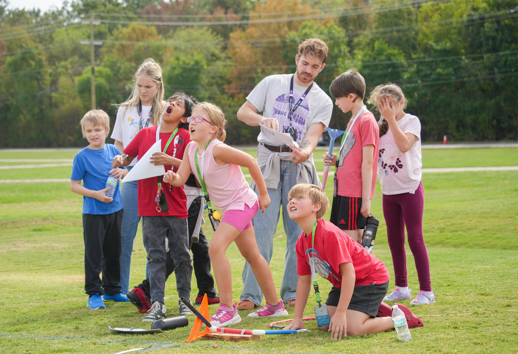 A group of children with a supervising adult, standing and kneeling on grass, holding paper flags.