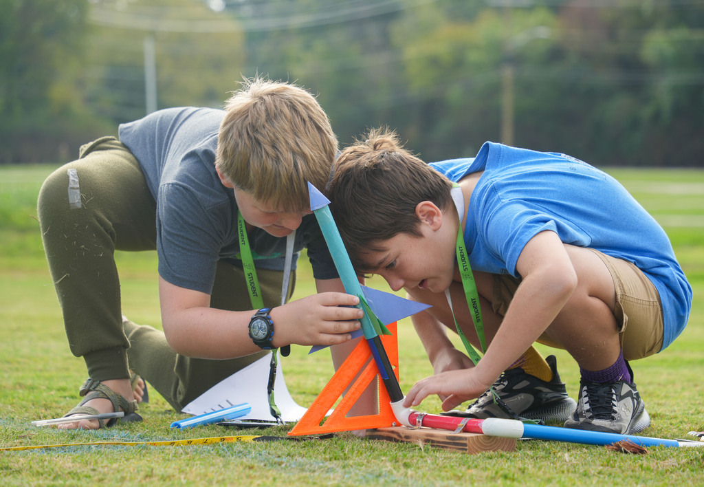 Two children crouch on a grass field, one holding a small rocket while the other inspects it.