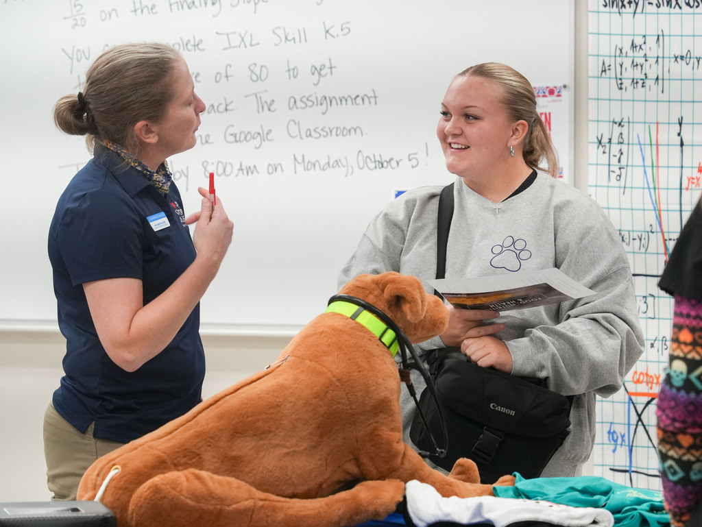 Two women and a dog in a classroom. One woman is holding a book and smiling. Another woman is writing on a whiteboard.