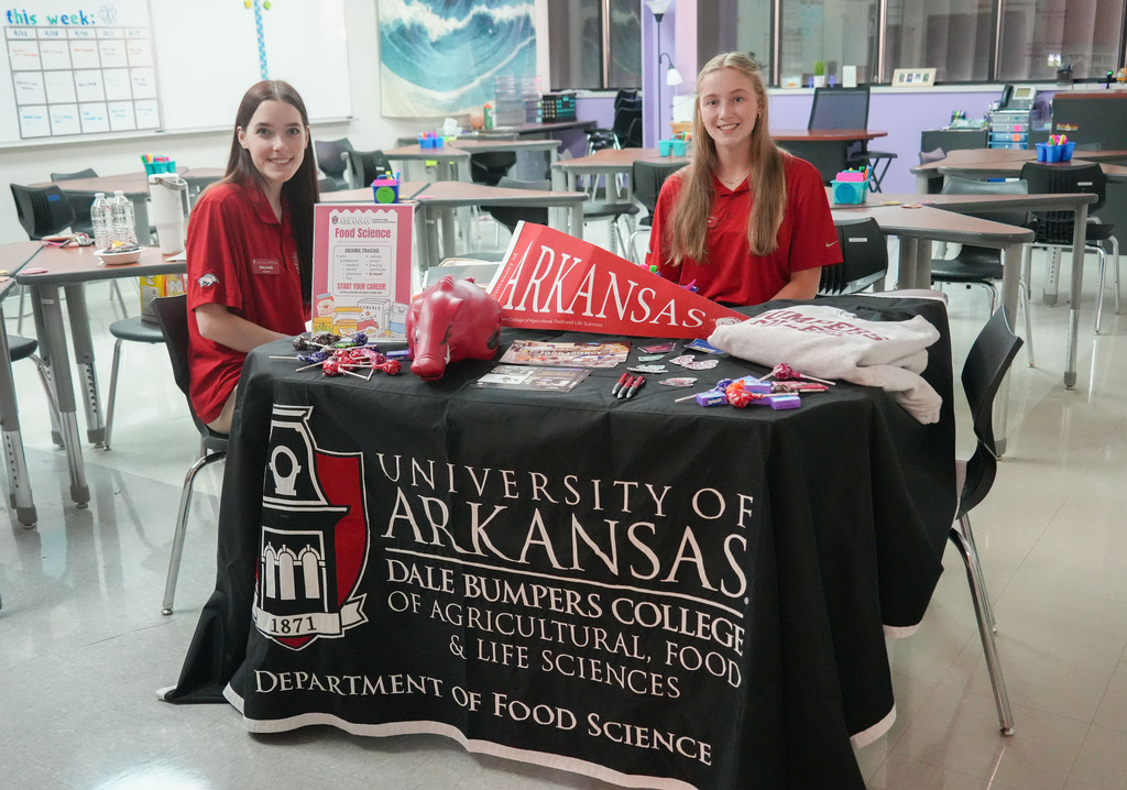 Two women in red shirts sit behind a table with University of Arkansas branding. Items are on the table.