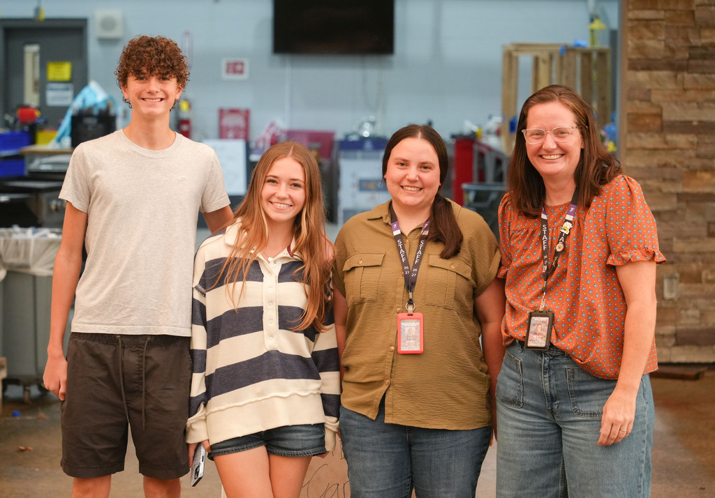 Four individuals stand close together, smiling, with a wall and objects behind them.
