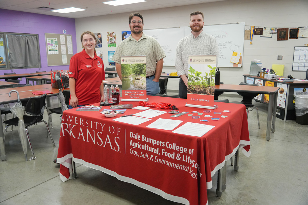 Three people stand behind a table. A red tablecloth has information on the table. White papers are on the table.