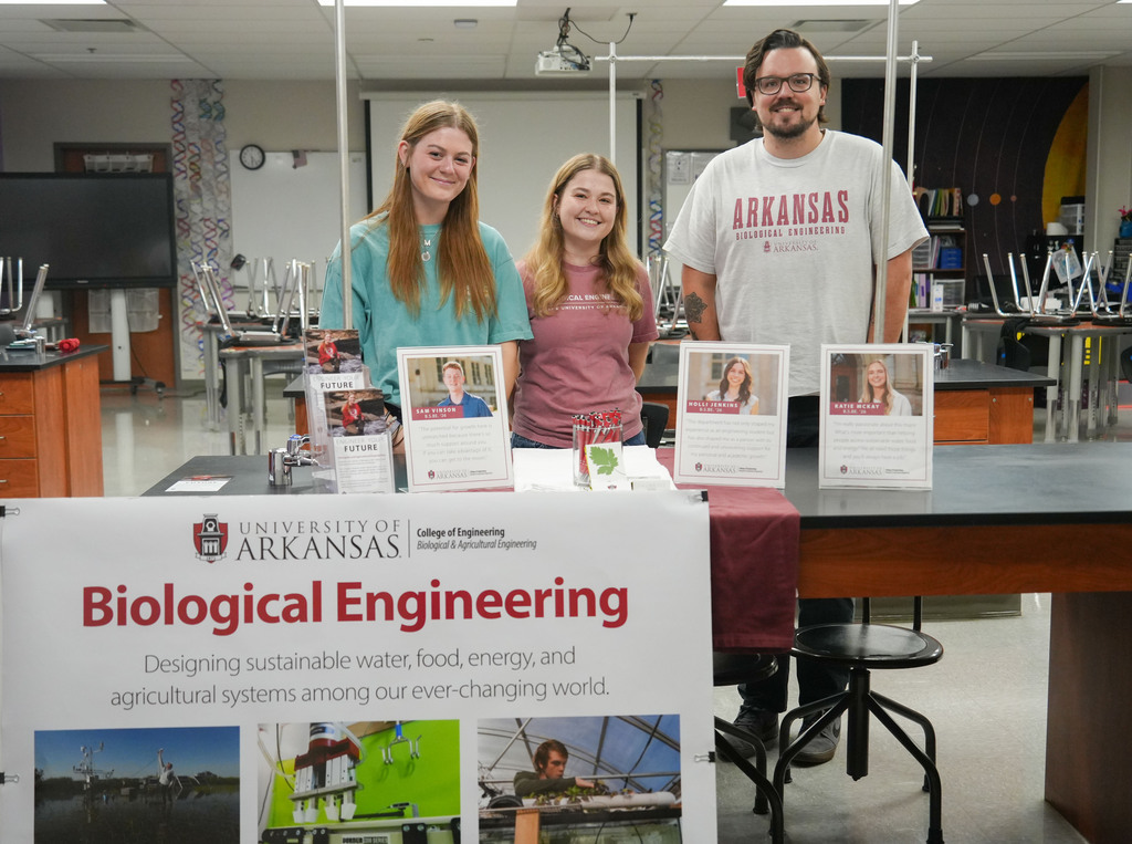 Three people pose in a lab. A banner reads "Biological Engineering" in front of them. The banner describes sustainable systems.