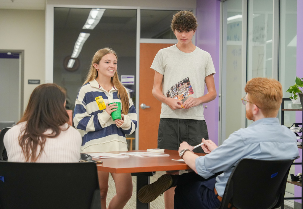 Four people sit or stand around a desk in an office; two hold cups and two look at papers.