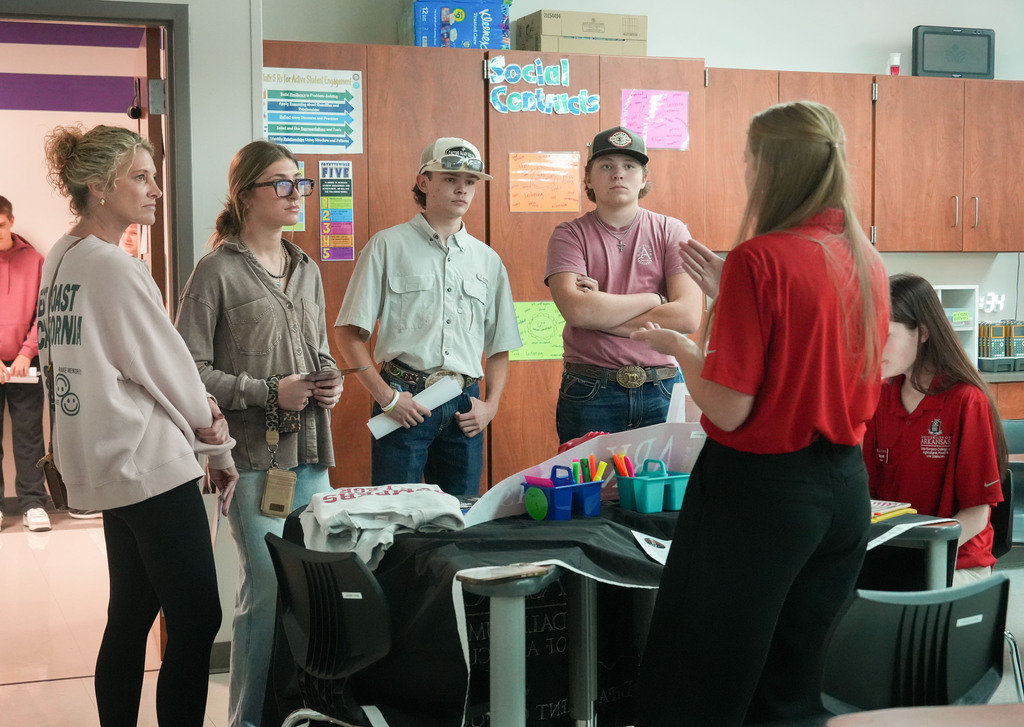 A group of people, including a woman speaking, stand around a desk in a classroom. Wooden cabinets and papers are in the background.