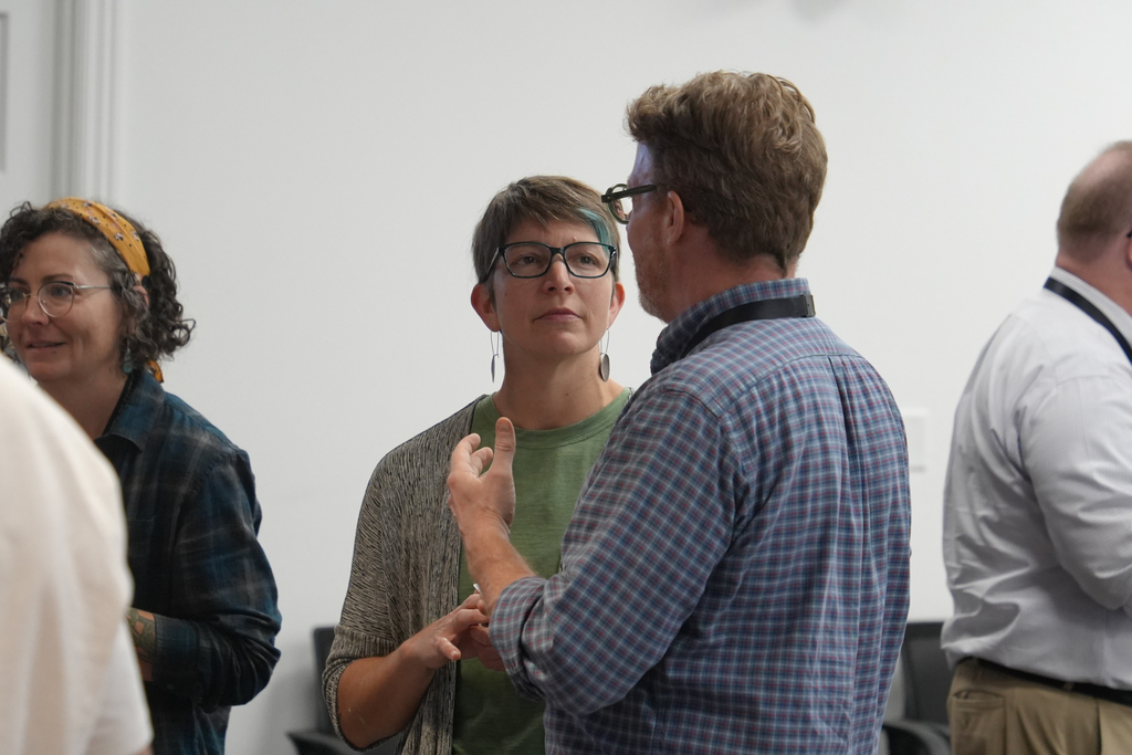A group of four adults in a room with white walls and chairs, two men and two women, engaged in conversation.