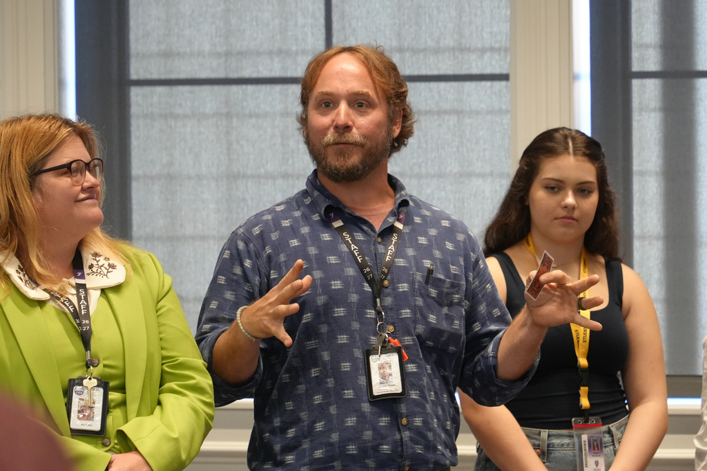 Three people stand in a room; one with glasses speaks with hands open. Behind them, a woman in a black top and another in a green jacket.