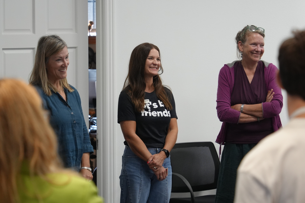 Four adults stand in a room, smiling. Two women face the group; one wears a black shirt with text, another wears a purple shirt.