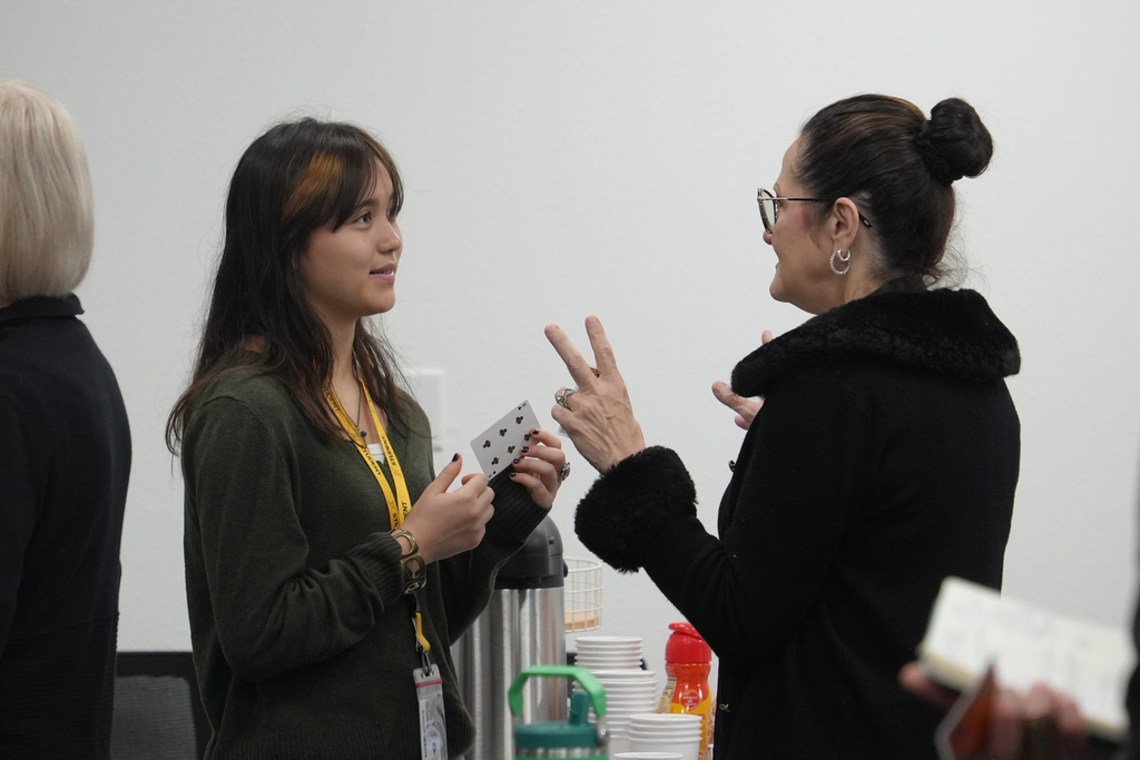 Two women converse, one holding playing cards. Behind them, a coffee station with cups and bottles.