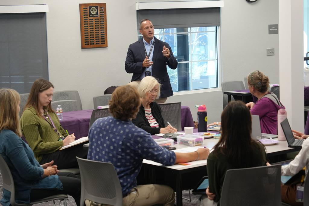 A man presents in a classroom. Several people sit at tables, listening and taking notes.