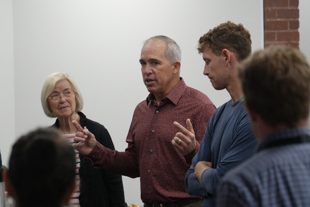 A man speaks to a group of people in a room; the man wears a maroon shirt, while the others wear blue and black.