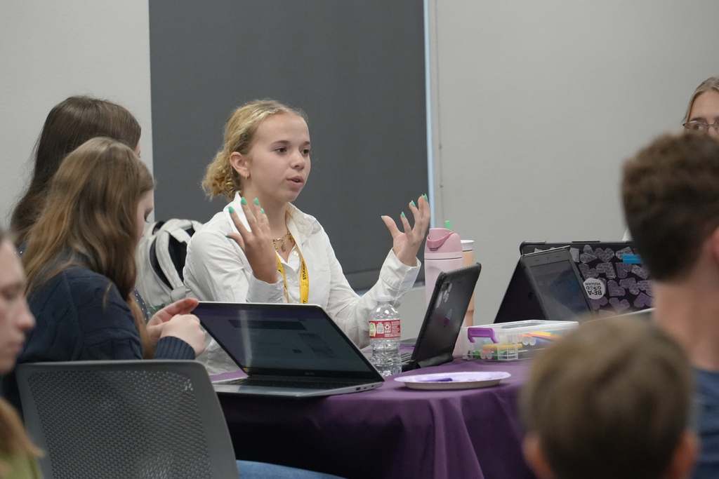 Several people seated around a table with laptops, one with green fingernails gesturing while talking.