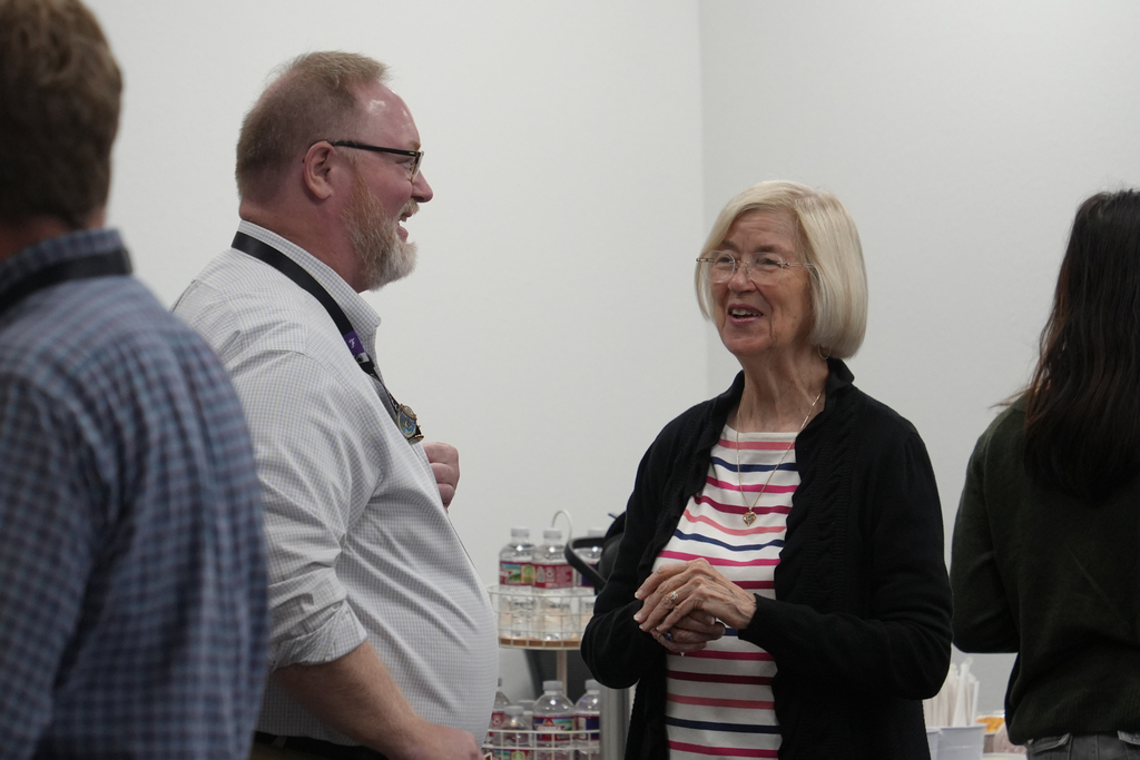 Three people converse in a white room with a shelf. One woman smiles, wearing glasses and a striped shirt. Two men stand, one wearing glasses and a stethoscope.