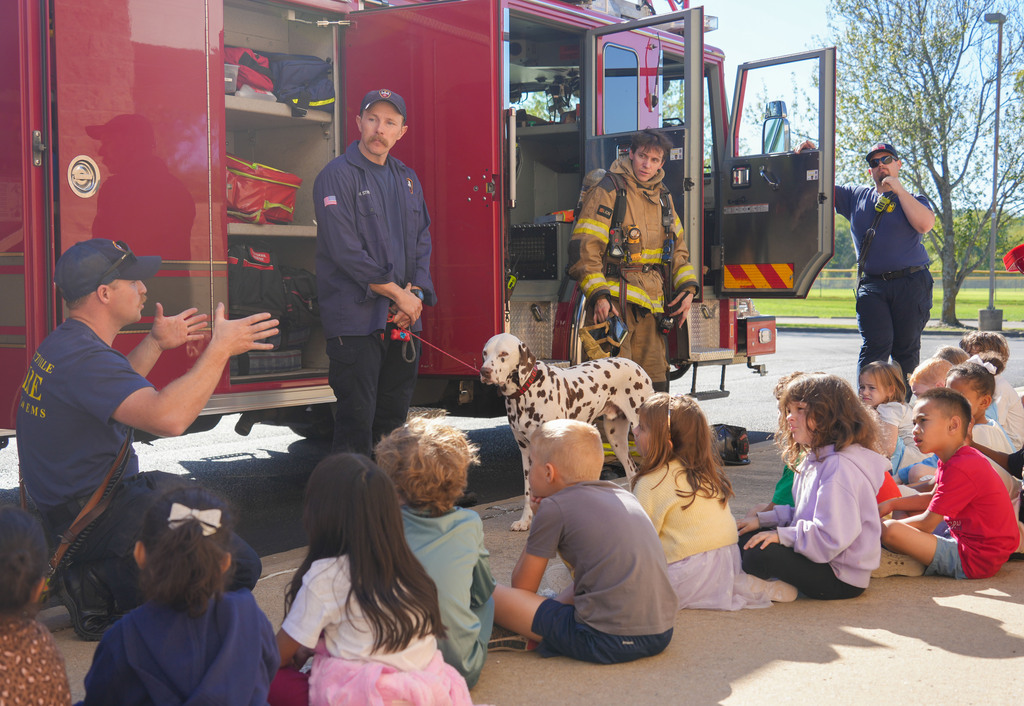 Children sit on the ground, listening to two firefighters and their dog. One firefighter holds the dog's leash.