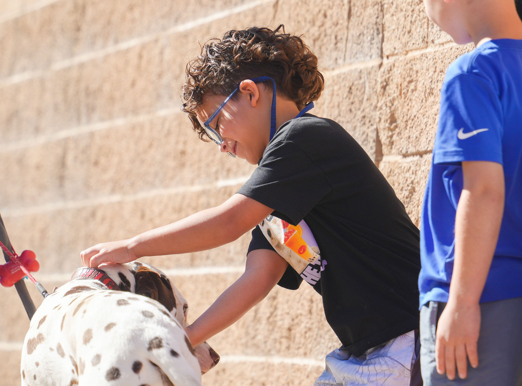 A child wearing glasses holds a dog's leash, and another child stands nearby against a brick wall.