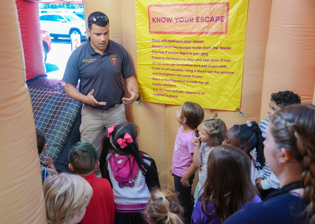 A firefighter speaks to a group of children in a bouncy house, with a yellow banner reading "KNOW YOUR ESCAPE".