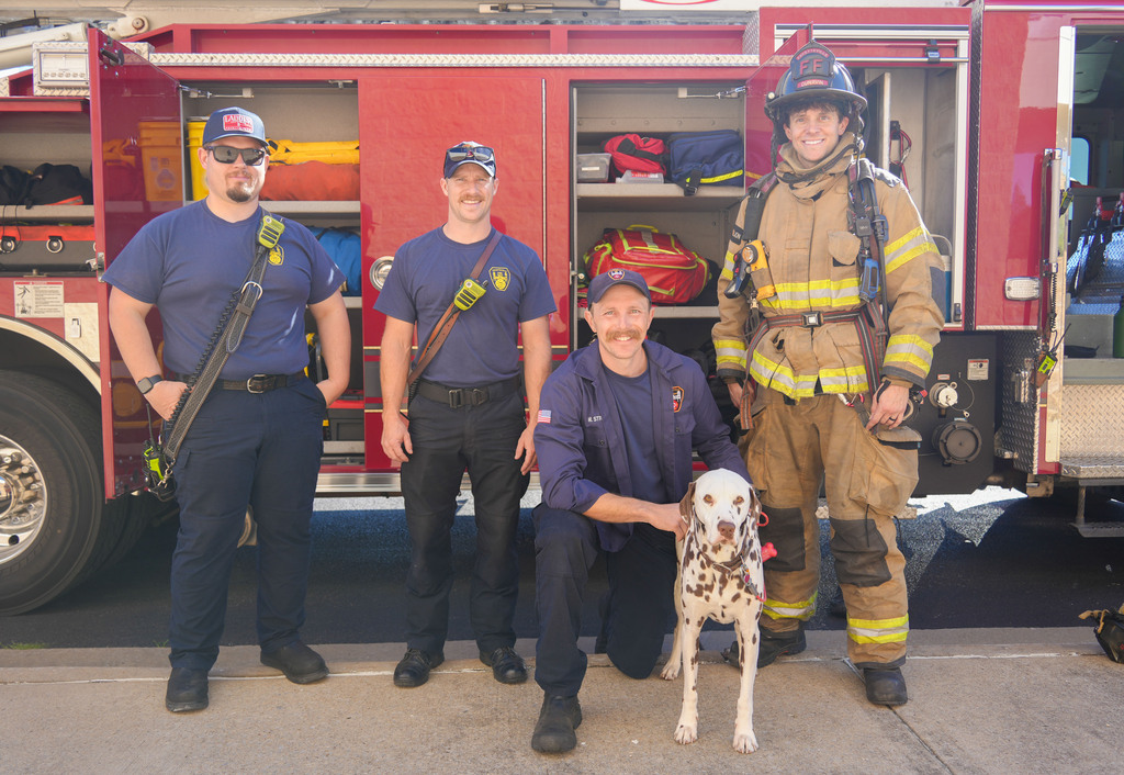 Five firefighters in blue uniforms and one in a full fireman's suit, stand in front of a firetruck. One kneels, interacting with a white and black spotted dog.