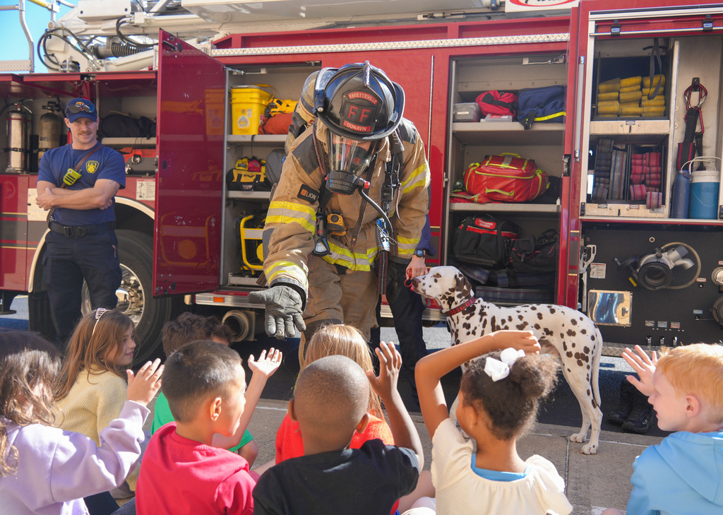 Fireman in protective gear, including a helmet and mask, stands before a group of children, a dalmatian beside him.