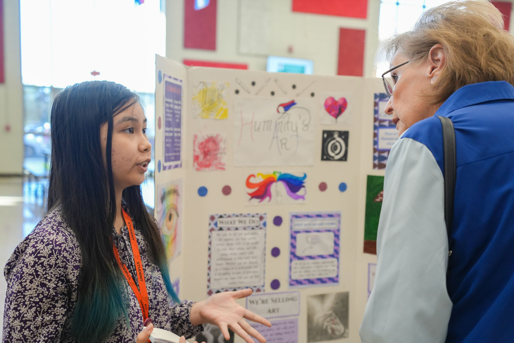 A girl speaks to an older woman in front of a colorful display with various drawings and writings.