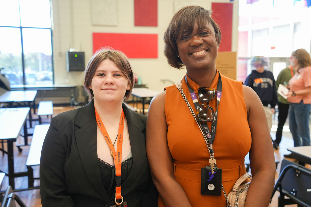 Two women stand next to each other, smiling. One wears a black blazer and the other an orange dress. They are in a room with desks and chairs.