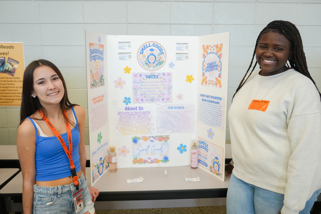 Two women stand before a table. One holds a presentation board displaying text and images. The other wears a white shirt.