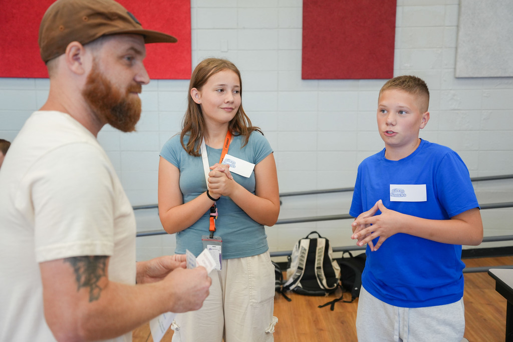 A man, a girl, and a boy in a classroom. The man wears a hat and white shirt. The girl in a blue shirt holds a paper.