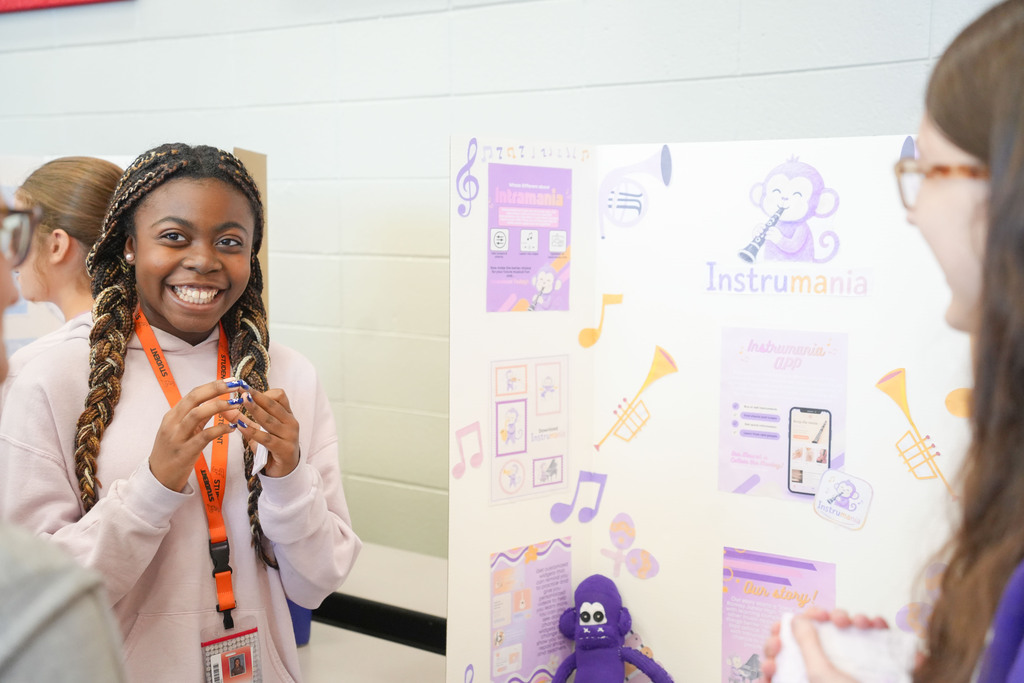 A woman with braided hair shows a small object to another woman with glasses. Behind them, a board displays musical instruments and a monkey.