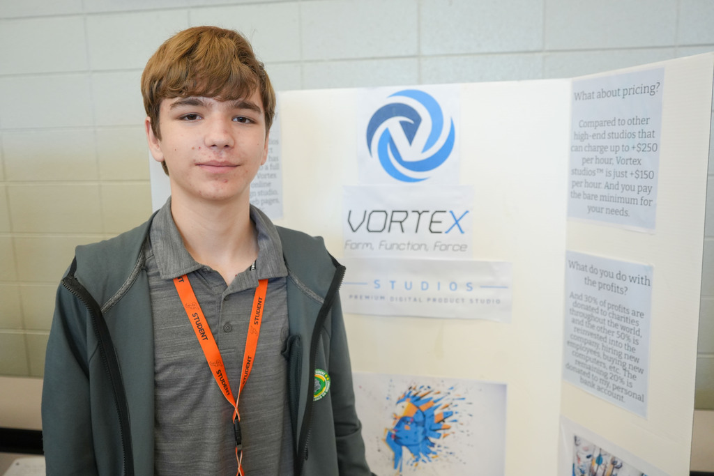 A boy stands in front of a Vortex display board, wearing a gray shirt and an orange lanyard.