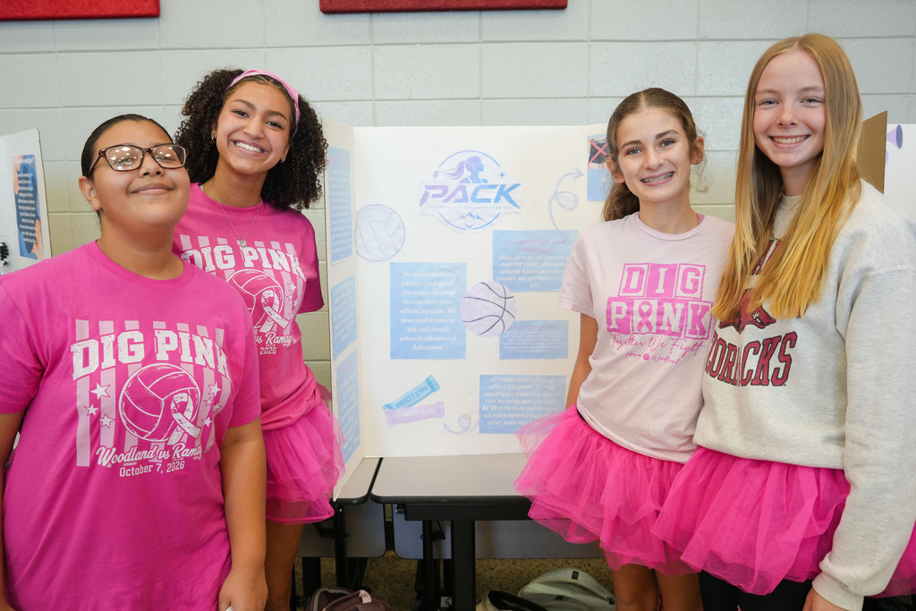 Four girls in pink shirts stand together in front of a poster. Two wear tutus, and the others wear glasses.