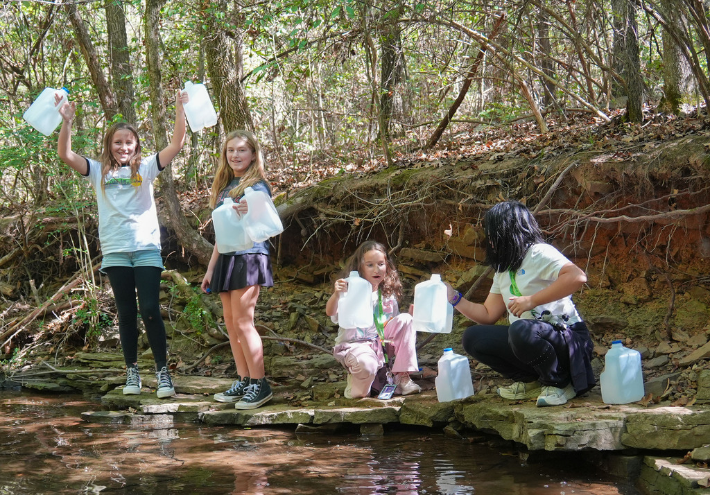 Four people hold plastic containers near a small stream. They stand on rocks. Trees and greenery surround them.