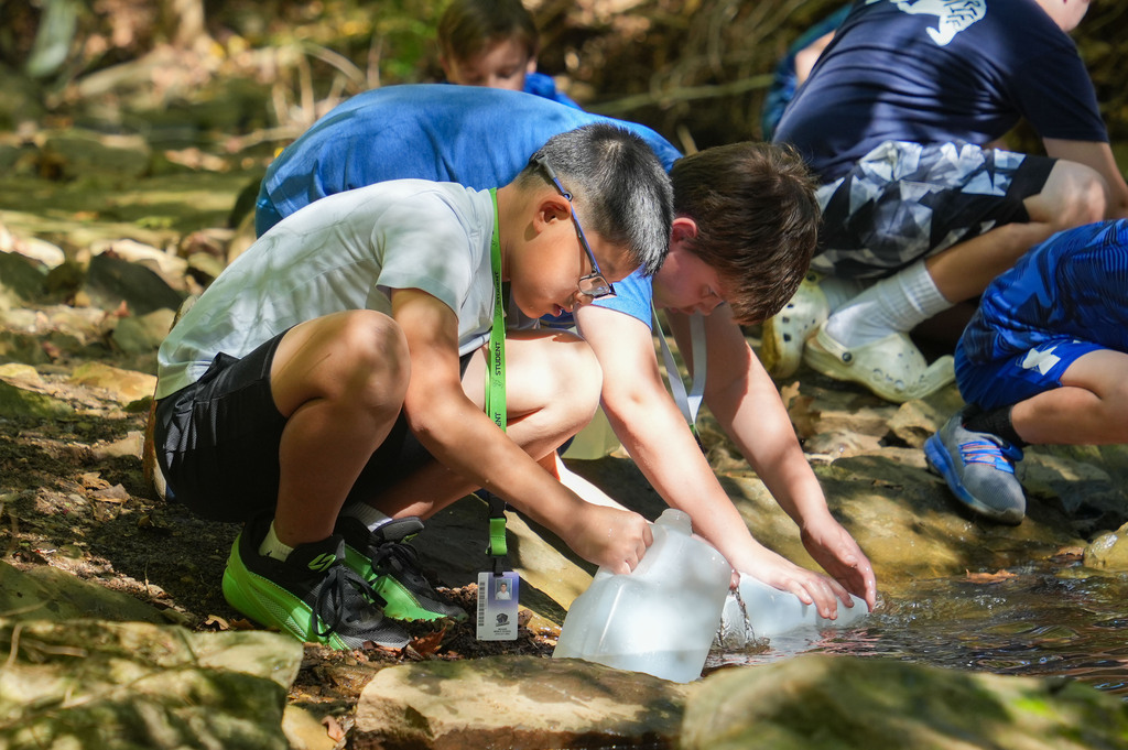 Kids examine water from a creek using equipment, with rocks and greenery in the background.