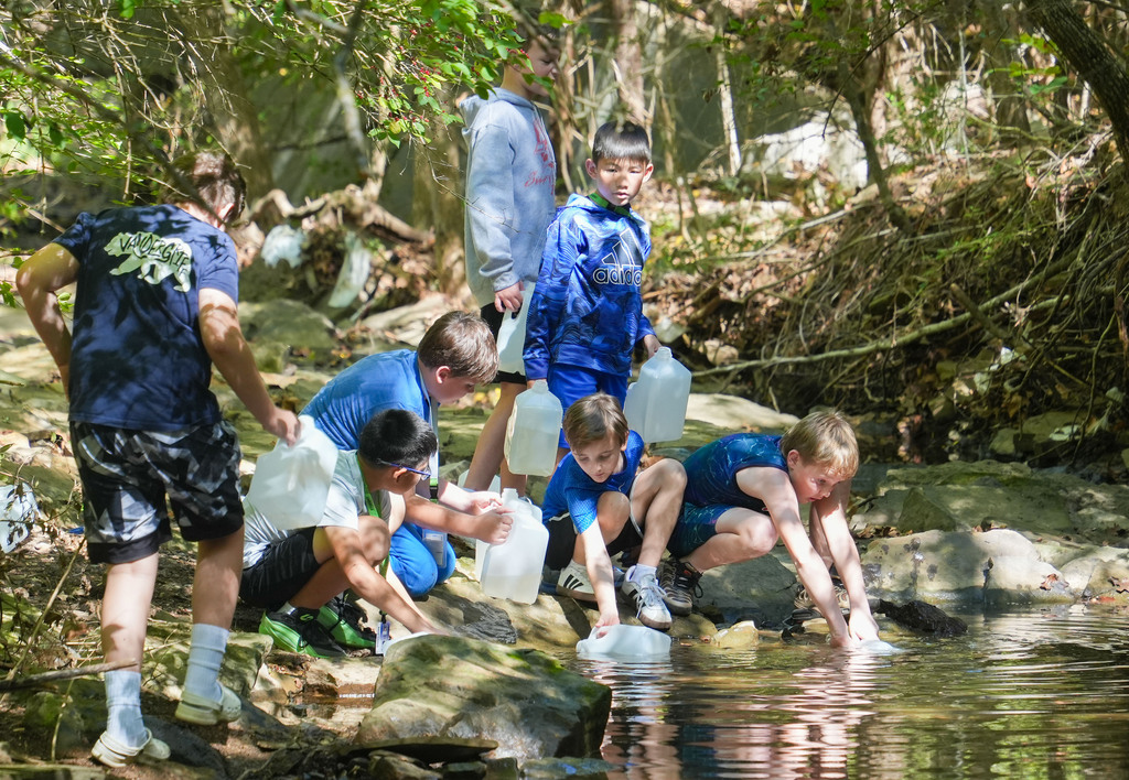 A group of children in blue uniforms collect water from a stream, some kneeling on rocks.