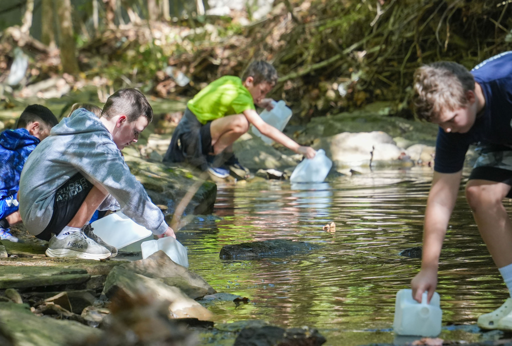 Four individuals near a creek, wearing casual attire. They are collecting water in plastic containers.