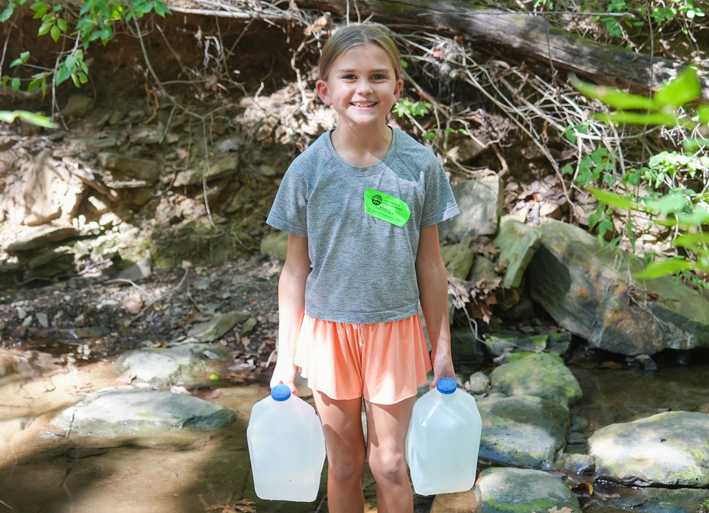 A child with a green sticker on their shirt holds two clear jugs in a rocky area.