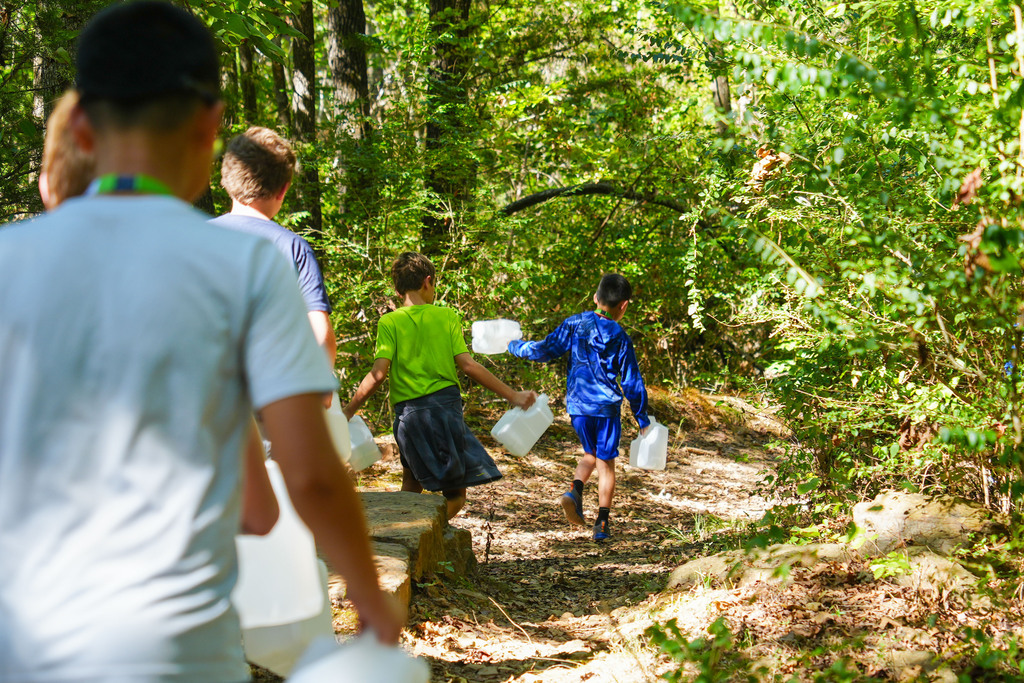 Group of people walking in a forest, carrying water jugs. Trees and plants surround the trail.