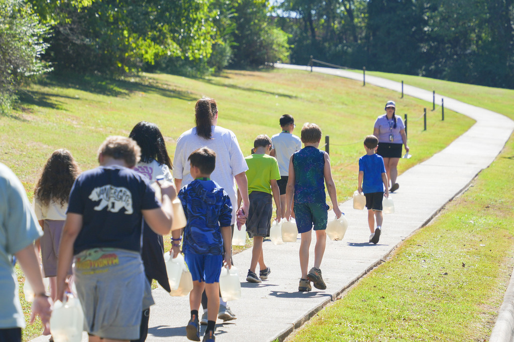 A group of people walk along a path carrying jugs, with a woman leading at the rear.