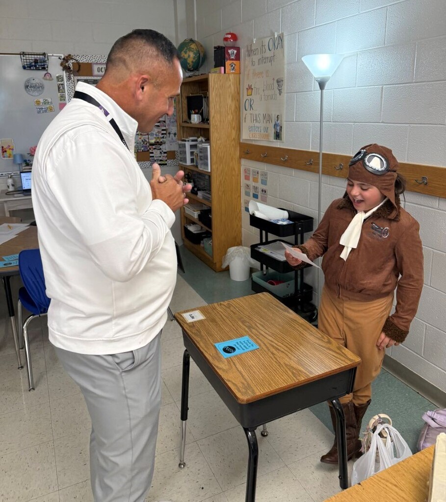 A man in a white shirt and a woman in a brown flight suit and goggles converse in a classroom.