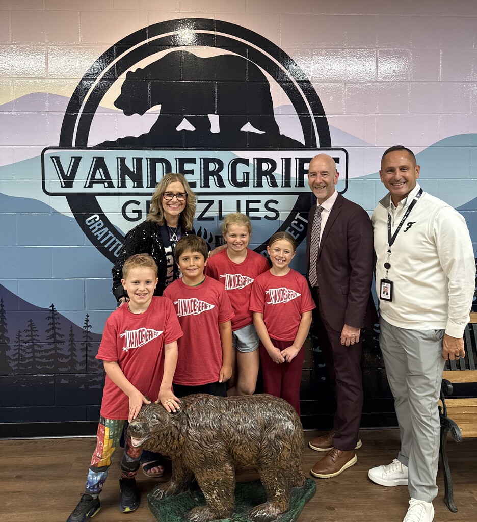 Four adults and six children pose with a bear statue. They wear red shirts with white writing. A wall behind them displays the words "VANDERGRIF GRIZZLIES" and a bear.