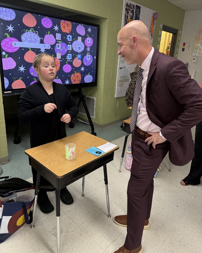 Man in a brown suit stands at a desk next to a young girl in a black jacket.