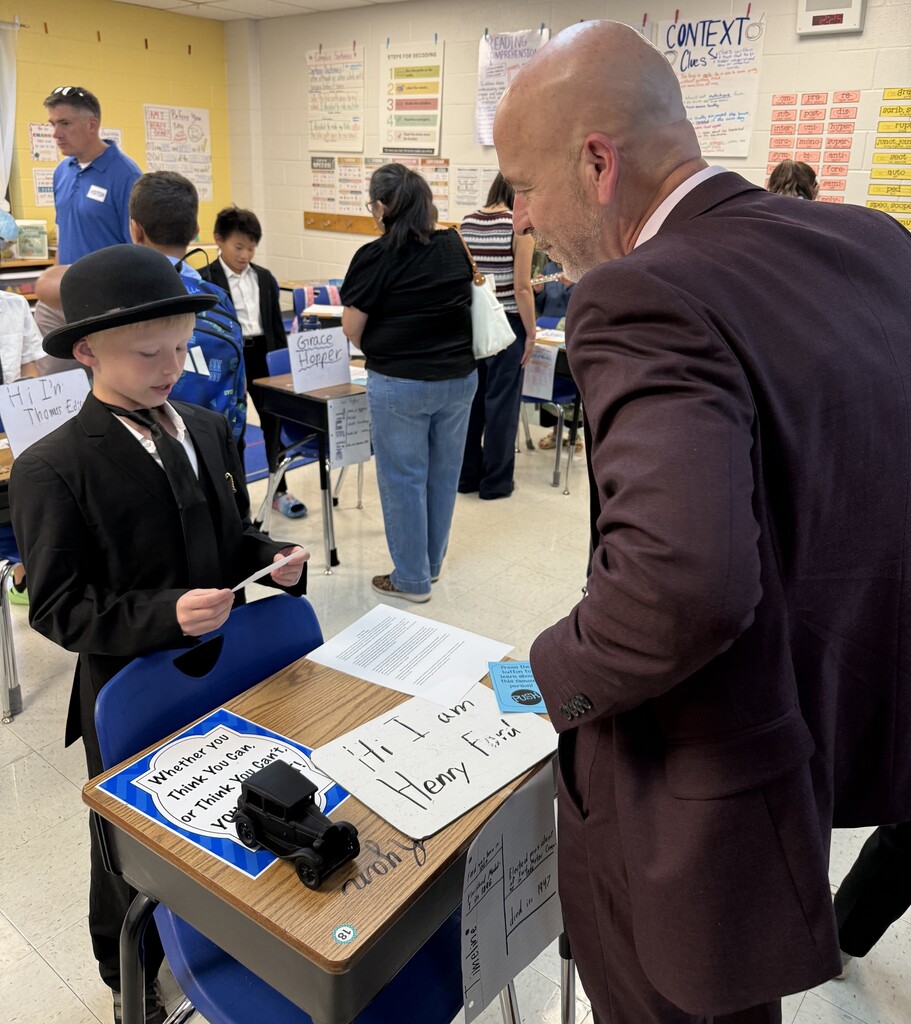 A man in a suit interacts with a child in a classroom setting. Behind them, more children and adults are present.