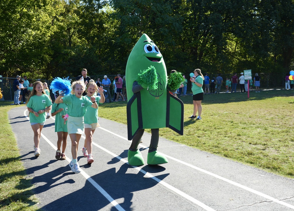 Children in green shirts run a track. A mascot in green holds green plants. People stand in the distance. Trees and grass surround the area.