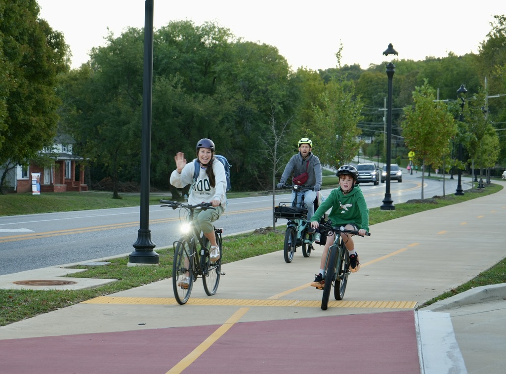Three people ride bicycles on a sidewalk. One wears a helmet and waves. Trees and streetlights flank the road.
