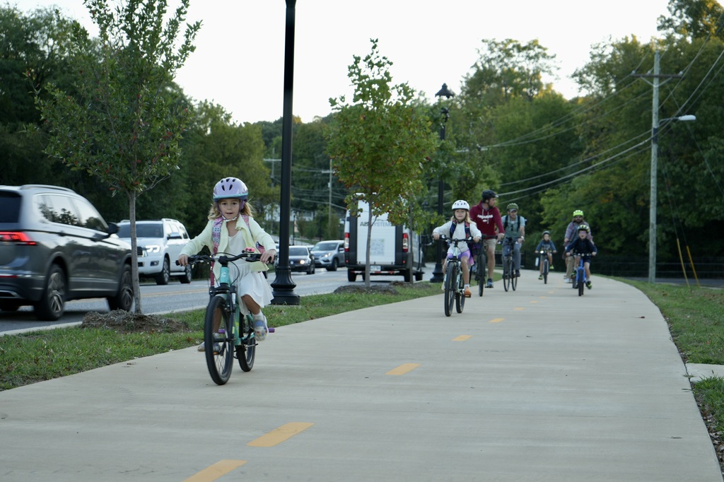 A group of people wearing helmets rides bicycles on a sidewalk with yellow markings. Trees and parked cars line the road.