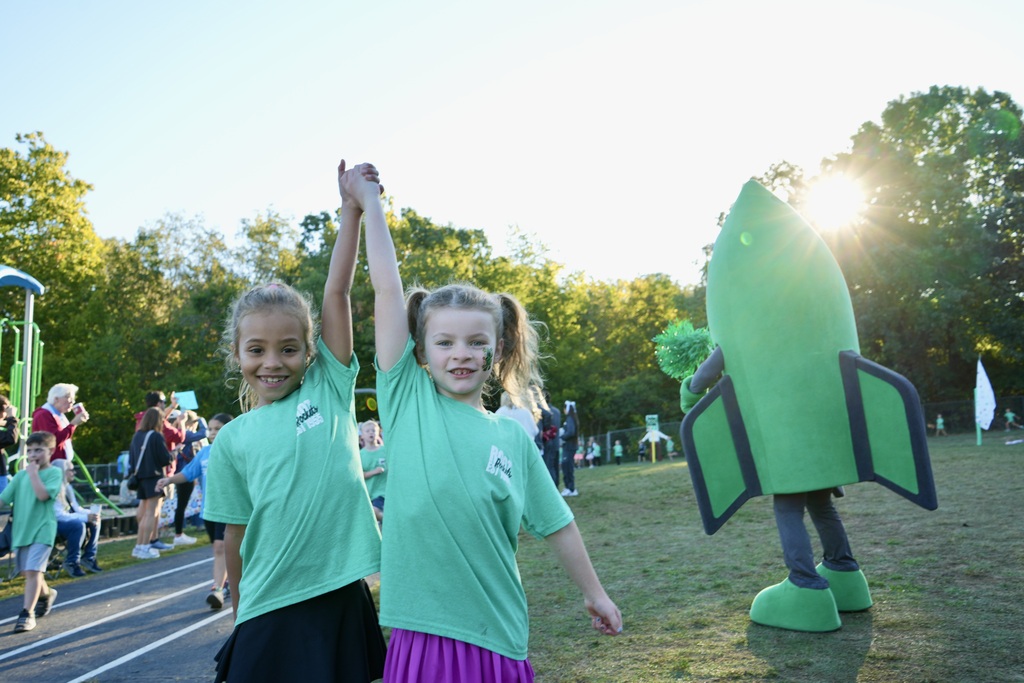 Two children in green shirts give high fives. A green rocket mascot stands behind them. Other people walk around, and trees and a playground are visible.