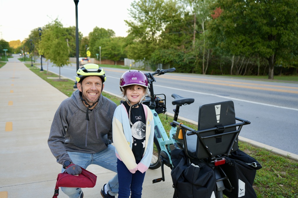 A man crouches, smiling, with a child beside him on a sidewalk. Both wear helmets. Bicycles with baskets and bags are nearby. Trees and a road are in the background.