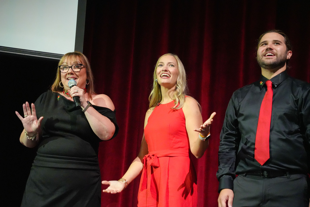 Three adults on a stage with a red curtain in the background, one woman speaks into a microphone.