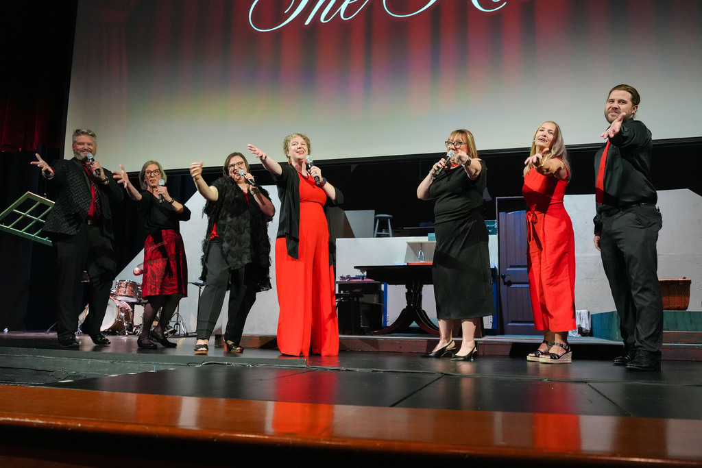 A group of people in red dresses, black suits, and high heels on stage, performing in front of a projection screen.