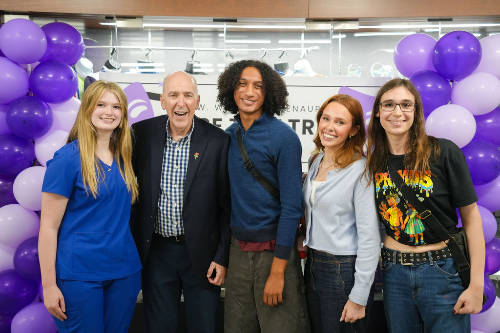 Five people stand together, smiling, in front of a backdrop of purple balloons.